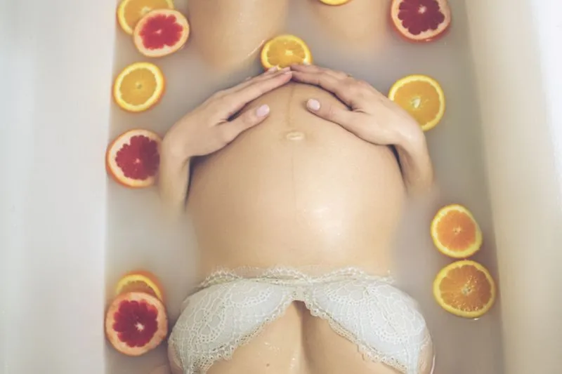 Femme enceinte allongée dans un bain laiteux, les mains sur le ventre, entourée de tranches d'orange et de pamplemousse