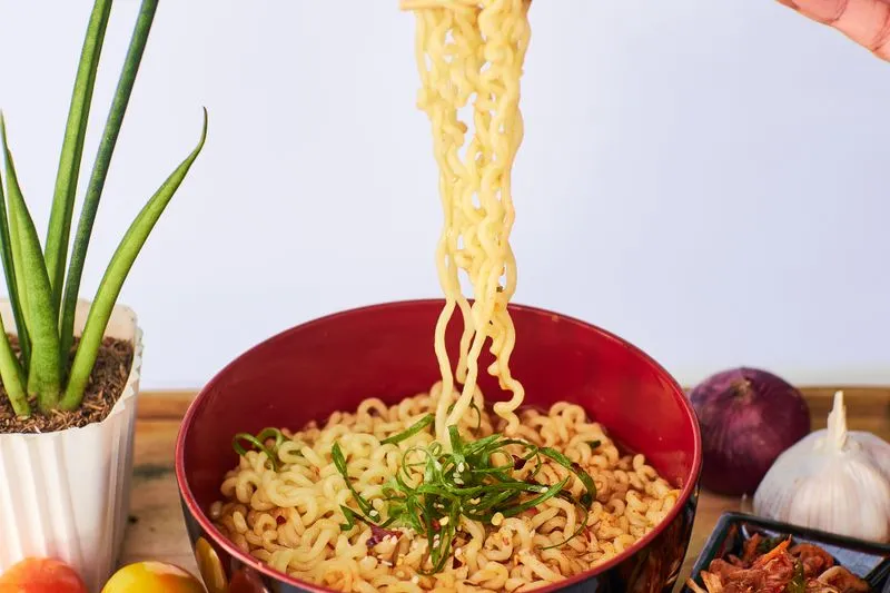 Nouilles instantanées soulevées avec des baguettes au-dessus d'un bol rouge, à côté d'une coupelle de kimchi