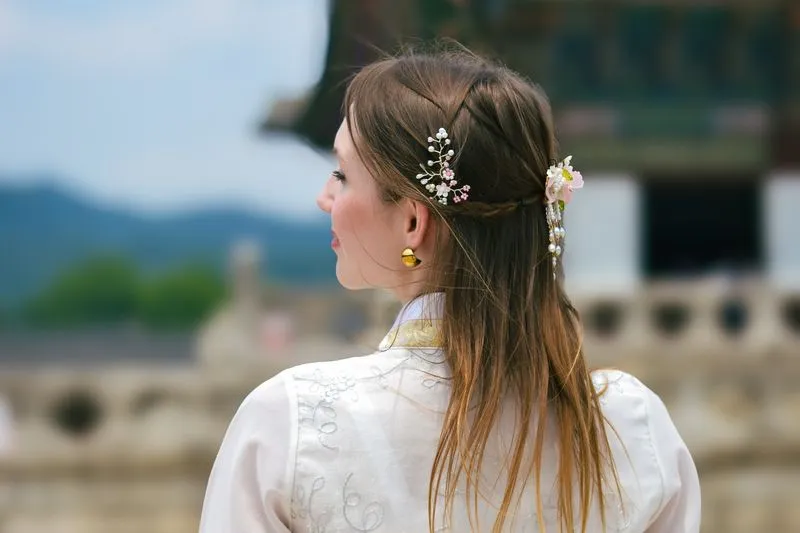 Jeune femme de dos en hanbok blanc brodé, des ornements fleuris dans les cheveux, devant un pavillon de palais coréen