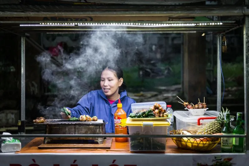 Vendeuse qui fait griller des brochettes derrière le comptoir de son stand de rue, au Vietnam
