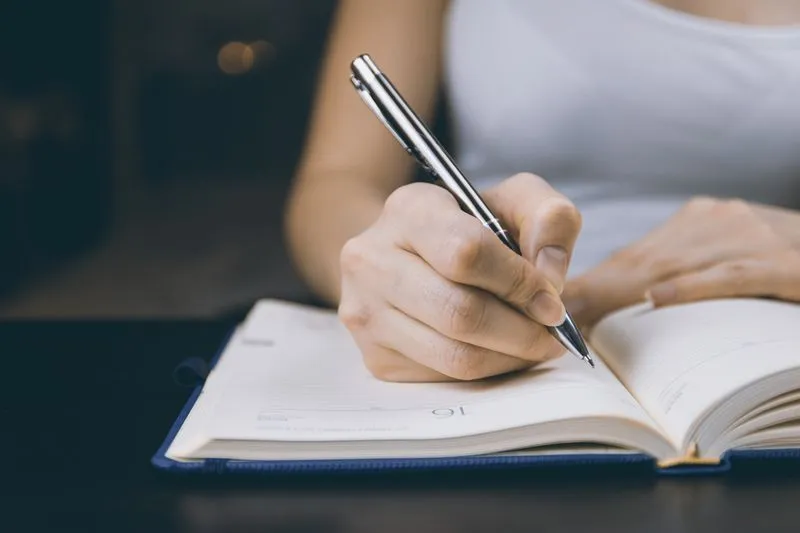Main qui écrit au stylo argenté dans un agenda ouvert à couverture bleu marine, posé sur une table sombre