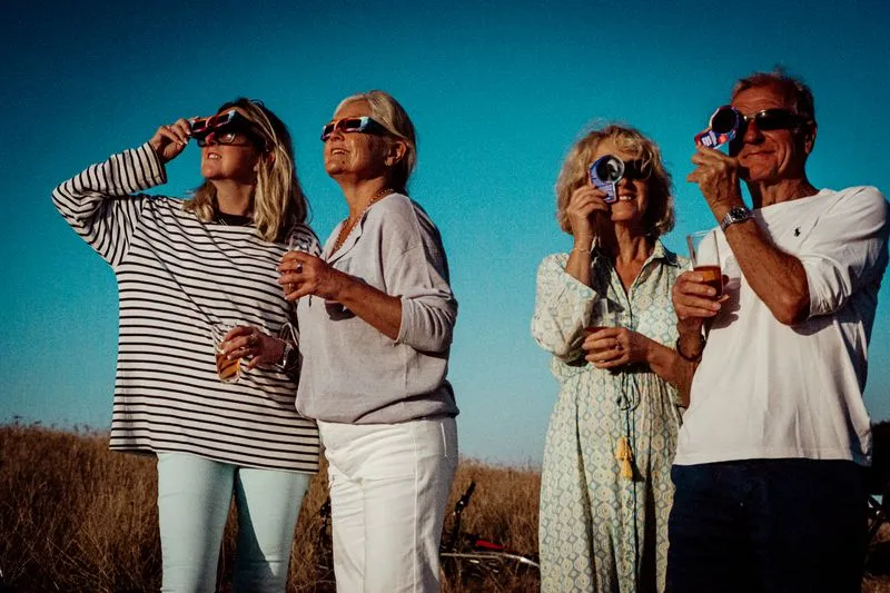 Quatre adultes en plein air, le visage tourné vers le ciel, qui observent une éclipse avec des lunettes et des visionneuses en carton