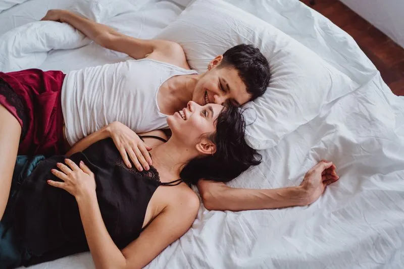 Couple allongé sur des draps blancs, qui rit, visage contre visage
