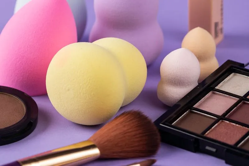 Éponges de maquillage de plusieurs formes et couleurs sur fond violet, avec un pinceau, une palette de fards et un fard compact