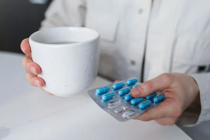 Mains qui tiennent un mug blanc et une plaquette de gélules bleues au-dessus d'une table