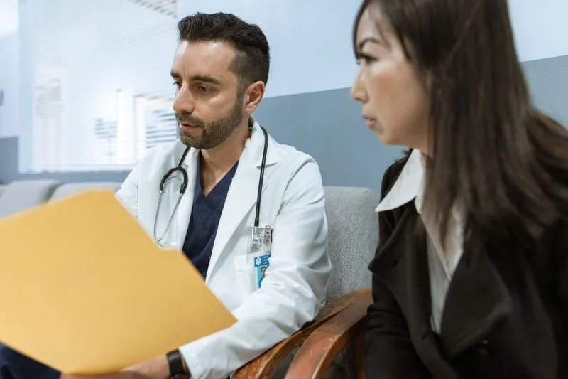 Médecin en blouse blanche, stéthoscope autour du cou, qui parcourt un dossier jaune assis à côté d'une patiente attentive