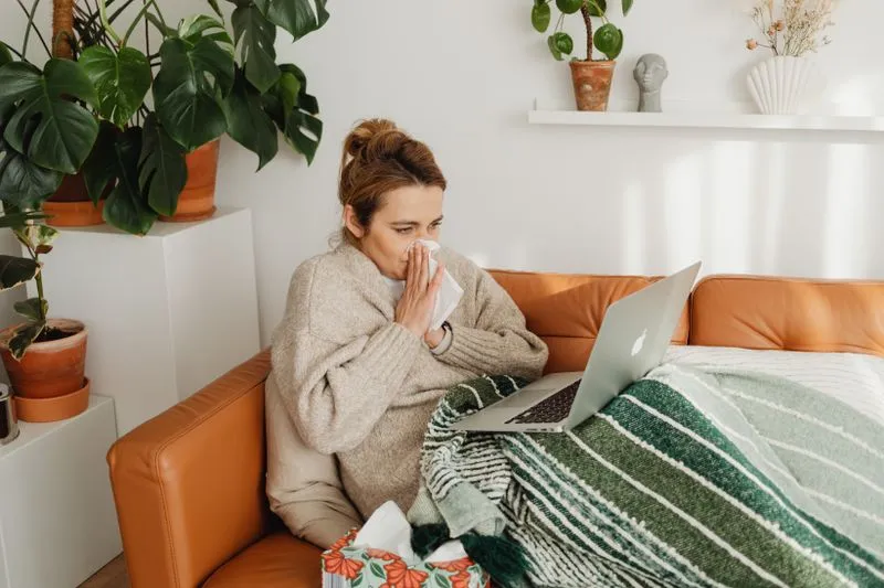 Femme installée sur un canapé sous un plaid, qui se mouche devant son ordinateur portable, une boîte de mouchoirs à ses pieds