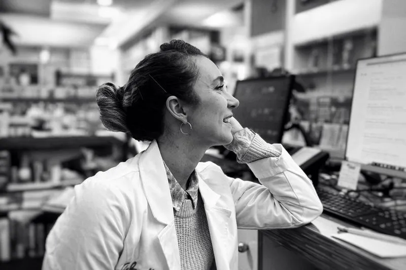 Pharmacienne souriante en blouse blanche, accoudée à son comptoir à côté de ses écrans d'ordinateur, photo en noir et blanc