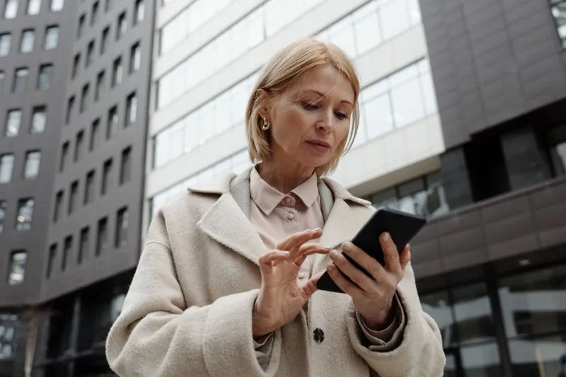 Femme en manteau clair qui consulte son smartphone dans une rue bordée d'immeubles