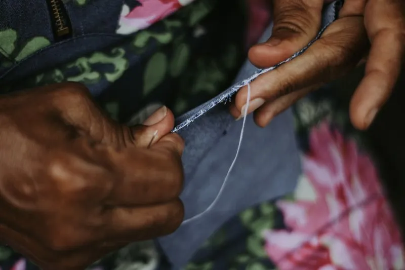 Mains qui cousent à l'aiguille le bord d'un tissu gris, un fil blanc tendu entre les doigts
