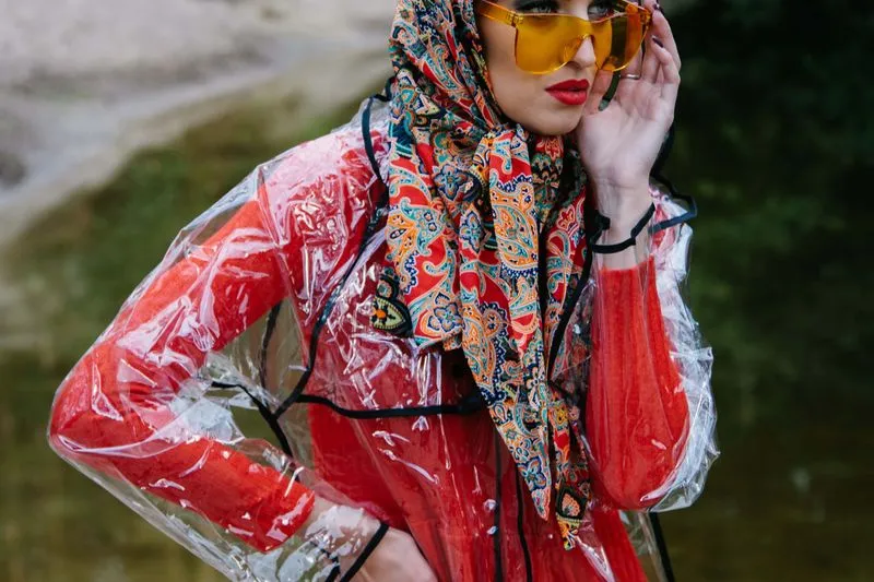 Femme coiffée d'un foulard imprimé cachemire, lunettes de soleil jaunes et imperméable transparent sur une robe rouge