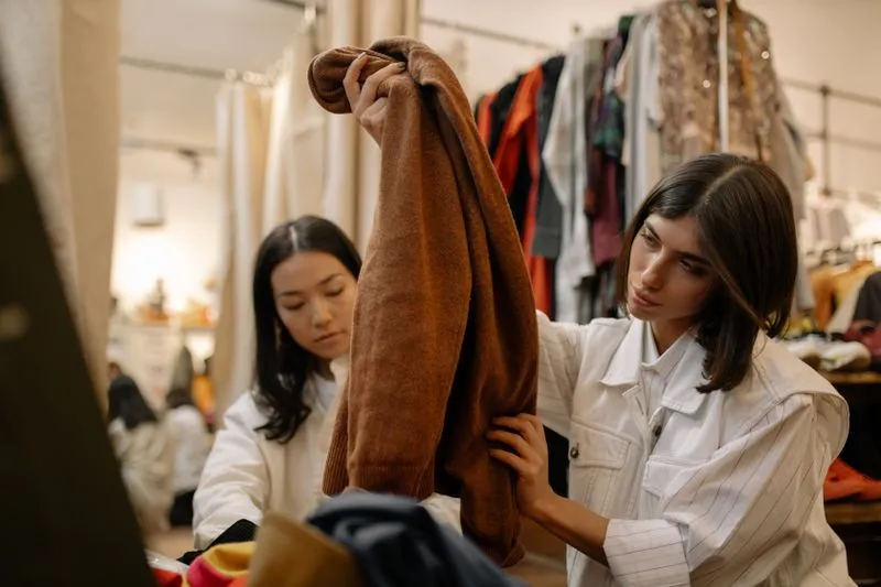 Deux femmes examinent un pull marron dans une friperie, devant des portants de vêtements