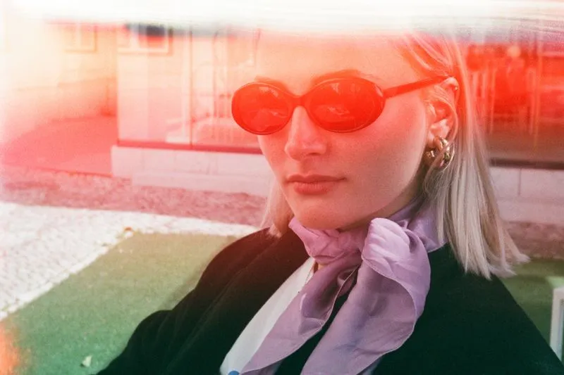 Femme blonde aux lunettes de soleil rouges, un foulard en soie lilas noué autour du cou sur une veste noire