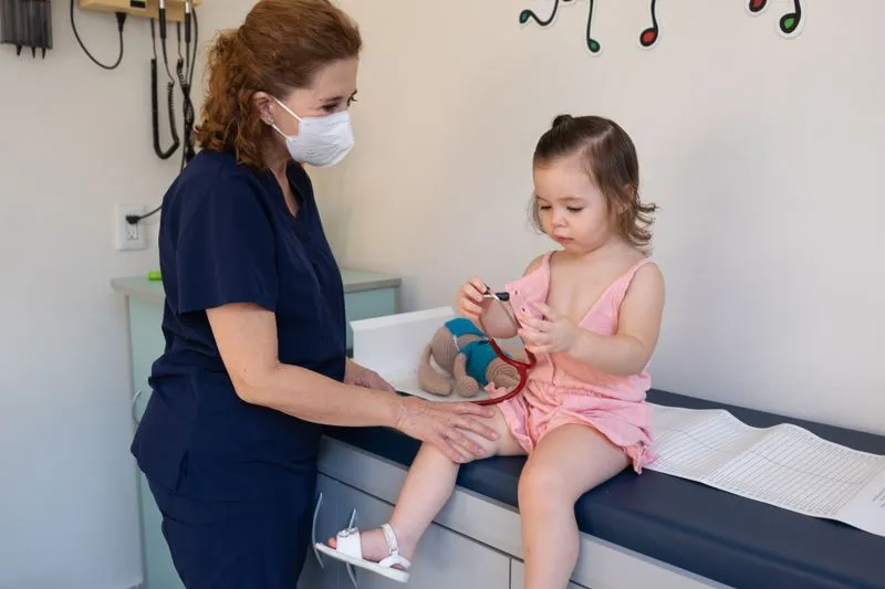 Fillette d'environ deux ans assise sur une table d'examen, qui manipule un stéthoscope à côté d'une médecin masquée, la main posée sur son genou