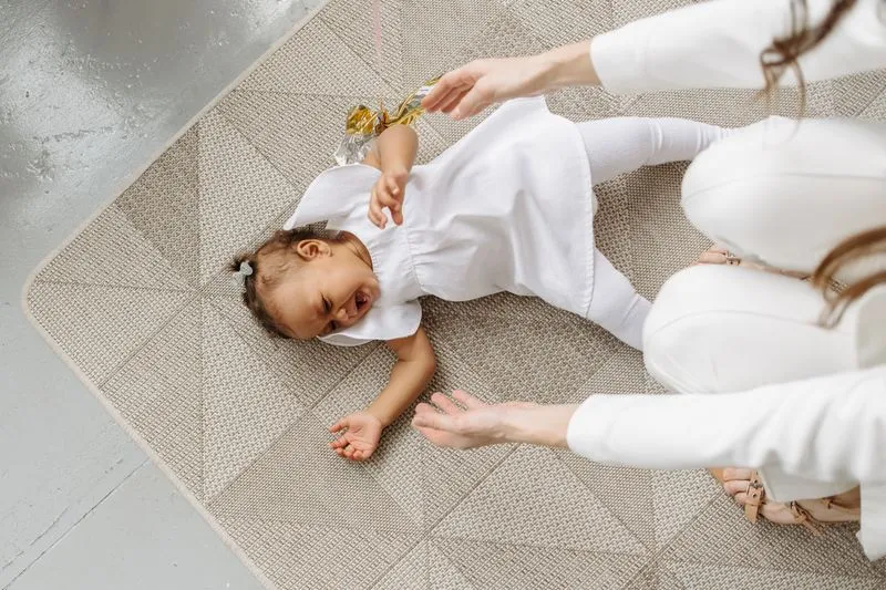 Petite fille allongée sur le dos sur un tapis, qui pleure en criant, pendant qu'un adulte accroupi tend les mains vers elle