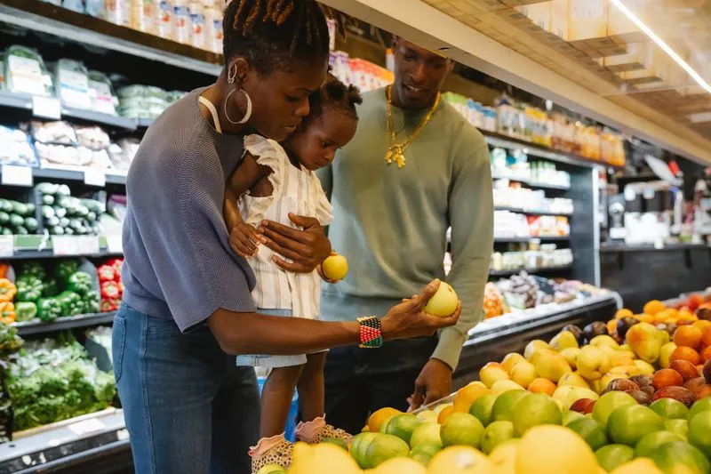 Mère qui porte sa petite fille sur la hanche devant l'étal de fruits d'un supermarché, l'enfant une pomme à la main, le père à côté