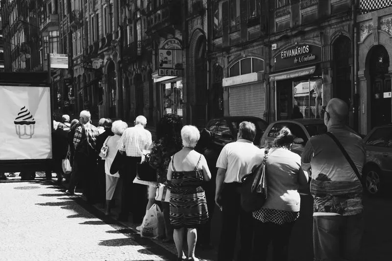 Photo en noir et blanc d'une longue file de personnes qui attendent sur un trottoir ensoleillé, devant des façades de boutiques anciennes