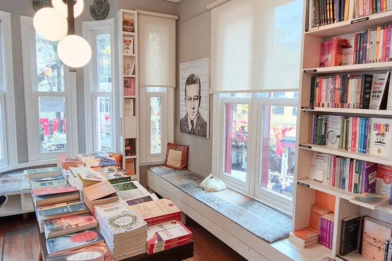 Coin d'une librairie lumineuse : table couverte de piles de livres, rayonnages blancs pleins et banquette sous les fenêtres