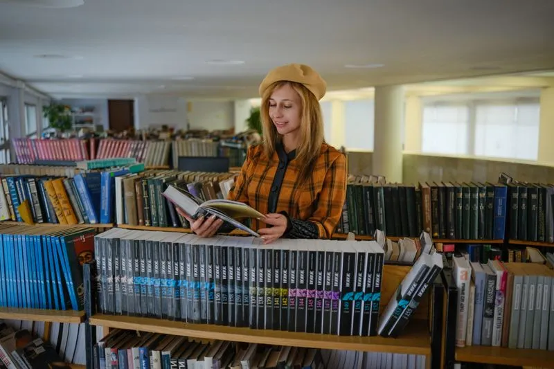 Lectrice en béret et veste à carreaux qui feuillette un livre ouvert entre des rayonnages remplis de livres