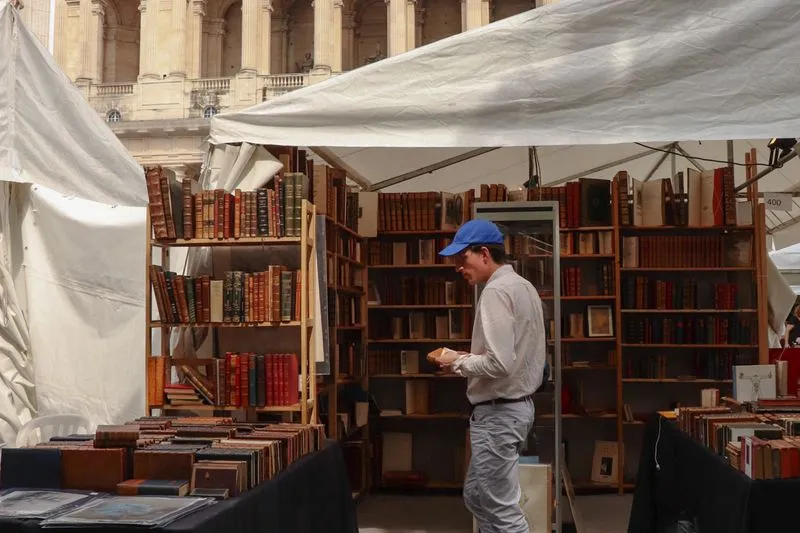 Stand sous une tente blanche dans un salon du livre en plein air, rayonnages de livres reliés et visiteur en casquette qui en feuillette un