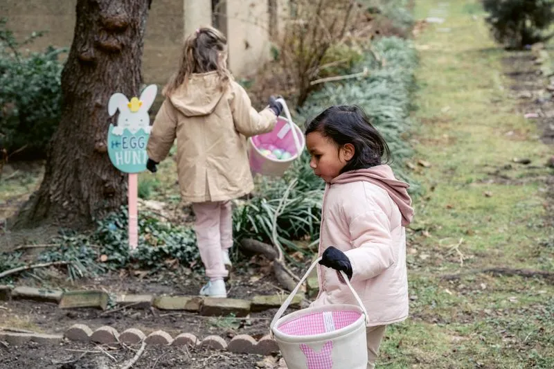 Deux petites filles en manteau, un panier de Pâques à la main, cherchent des œufs dans un jardin près d'un panneau « Egg hunt »