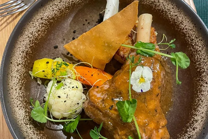 Souris d'agneau braisée nappée de son jus, servie avec des carottes, du chou-fleur et des pousses de pois