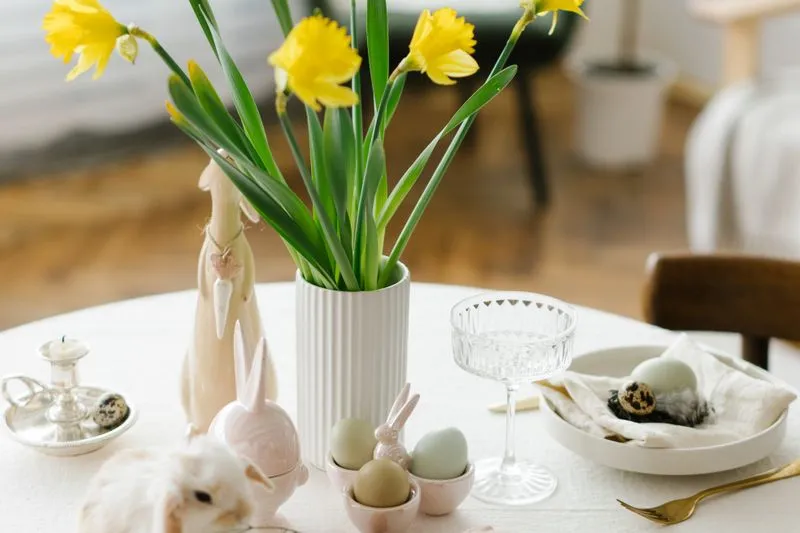 Table de Pâques dressée avec un vase de jonquilles, des œufs pastel dans des coquetiers, des lapins en céramique et un petit lapin au premier plan