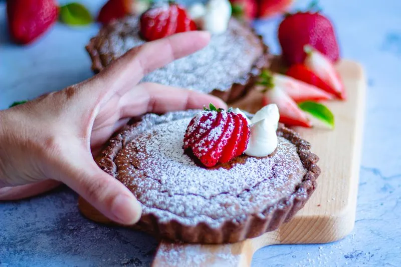 Tartelettes au chocolat saupoudrées de sucre glace, garnies d'une fraise et de crème fouettée, sur une planche en bois