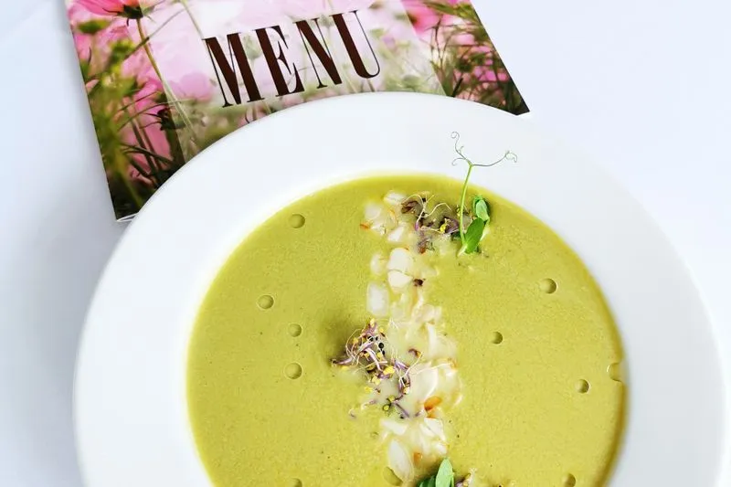 Velouté de petits pois dans une assiette creuse blanche, garni de pousses de pois et de graines germées, à côté d'une carte de menu fleurie