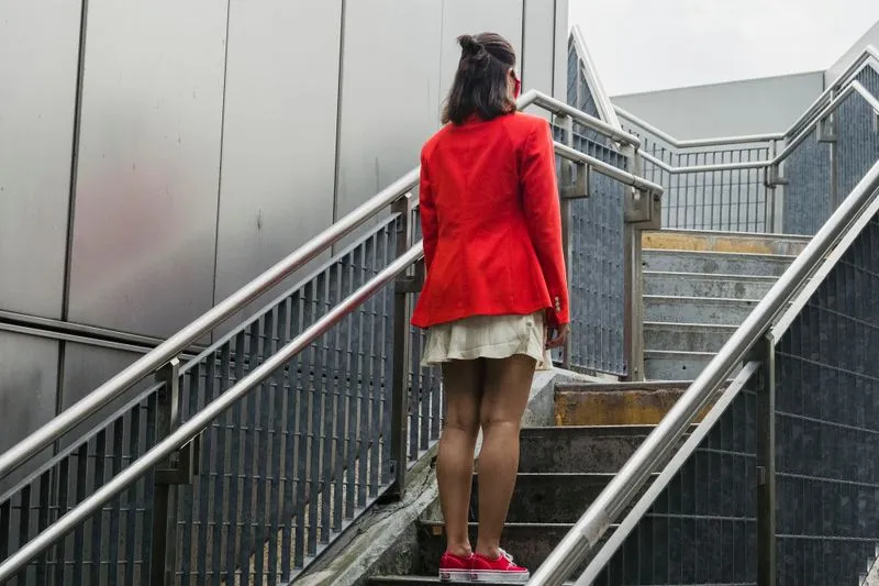 Jeune femme en veste rouge et baskets rouges, vue de dos, au pied d'un escalier en béton en ville