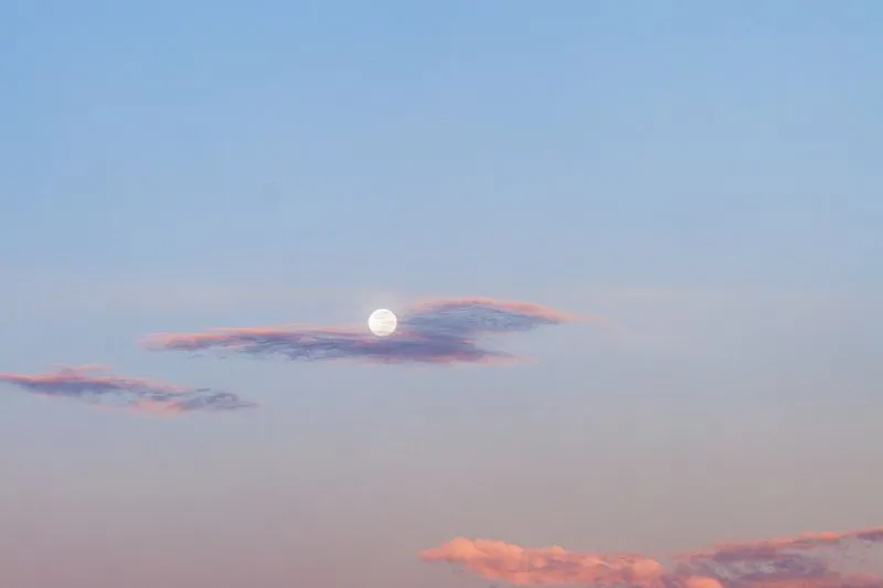 Pleine lune blanche entre deux nuages roses, dans un ciel bleu pâle au crépuscule