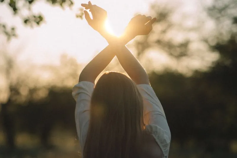 Jeune femme aux longs cheveux, vue de dos, bras levés et croisés vers un soleil rasant qui filtre entre les arbres