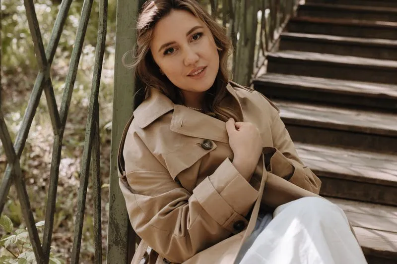 Femme assise sur des marches en bois, en trench beige court : col à revers, rabat de poitrine boutonné et pattes de poignet