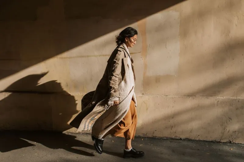 Femme qui marche le long d'un mur ensoleillé en trench beige long et ouvert, sur une jupe midi rouille et des derbies noirs