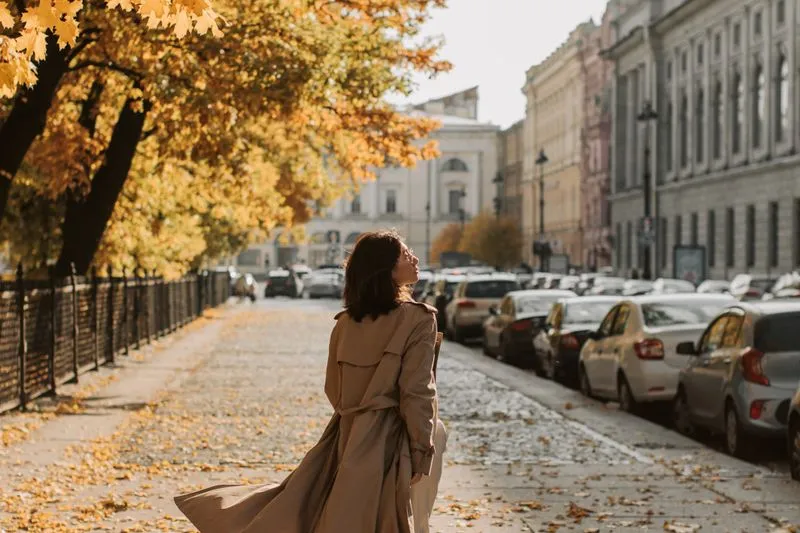 Femme de dos en trench beige ceinturé, pans soulevés par le vent, sur une avenue pavée bordée d'immeubles anciens et d'arbres aux feuilles jaunes d'automne