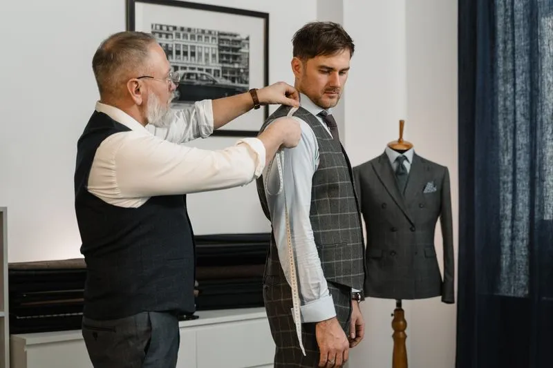 Tailleur qui mesure au mètre ruban l'épaule d'un client en gilet, une veste de costume sur un mannequin derrière eux