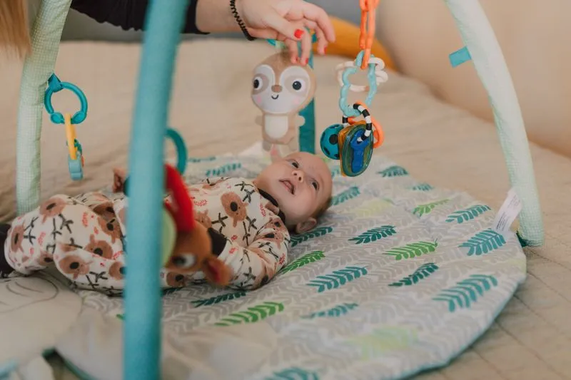 Bébé allongé sur le dos sous une arche d'éveil, qui sourit aux jouets suspendus qu'une main d'adulte fait bouger