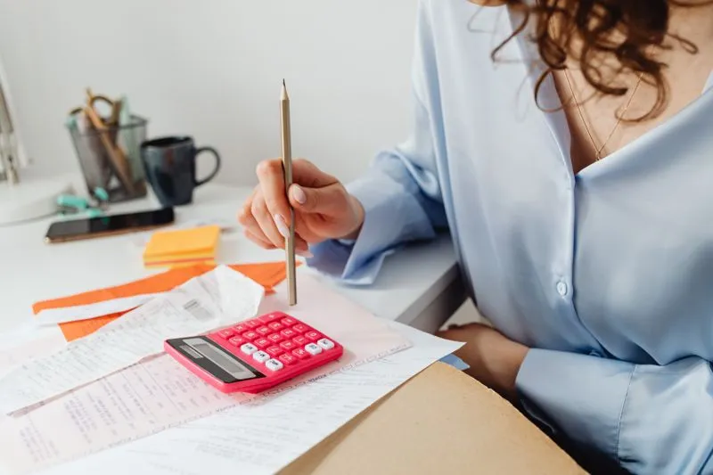Femme en chemisier bleu clair, crayon à la main, devant une calculatrice rose posée sur des factures et des tickets