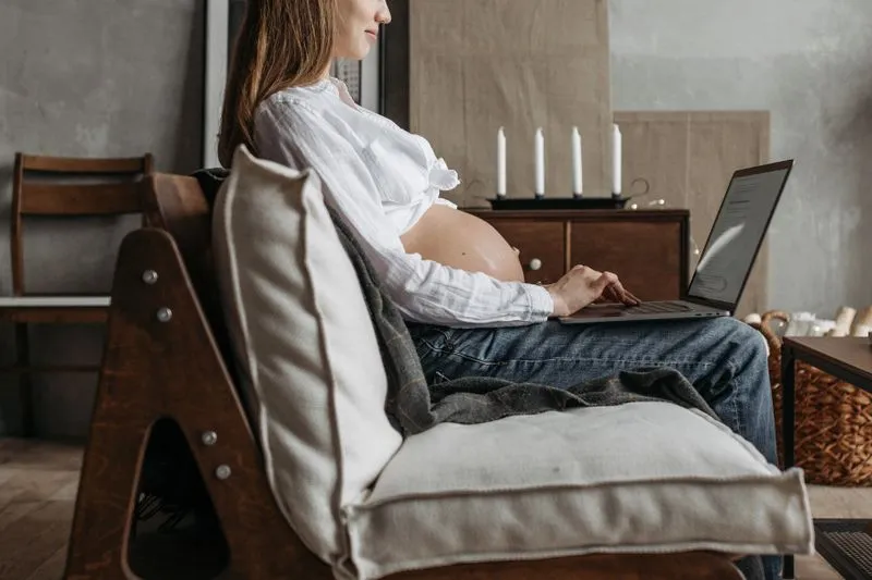 Femme enceinte assise dans un fauteuil en bois qui tape sur un ordinateur portable posé sur ses genoux