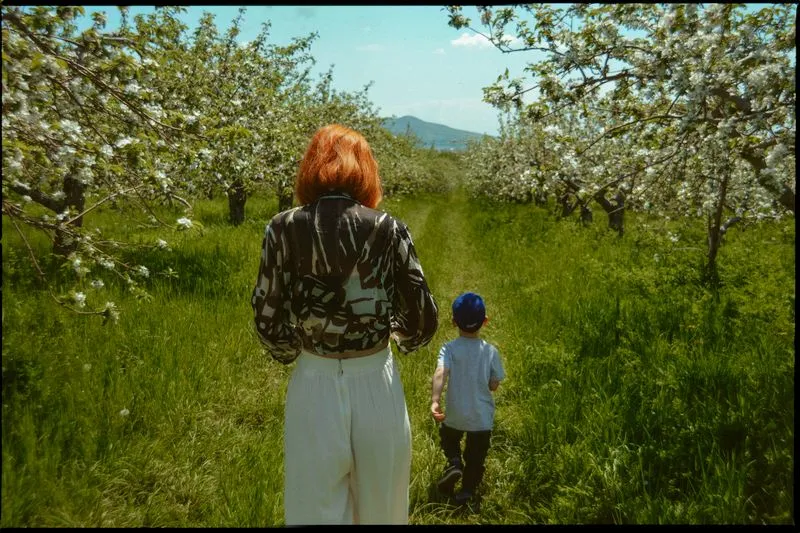 Femme rousse qui marche derrière un petit garçon en casquette dans un verger de pommiers en fleurs