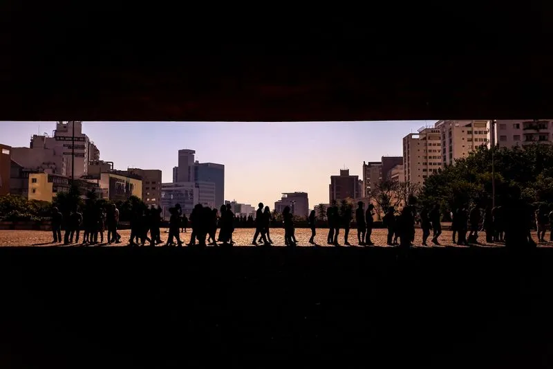 Silhouettes d'une longue file de personnes qui attendent sous une grande dalle de béton, avec des immeubles en arrière-plan