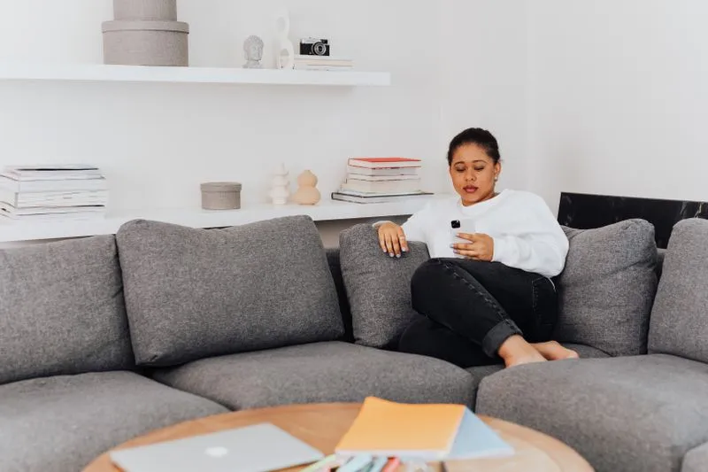 Jeune femme en pull blanc assise sur un canapé gris, les yeux sur son smartphone