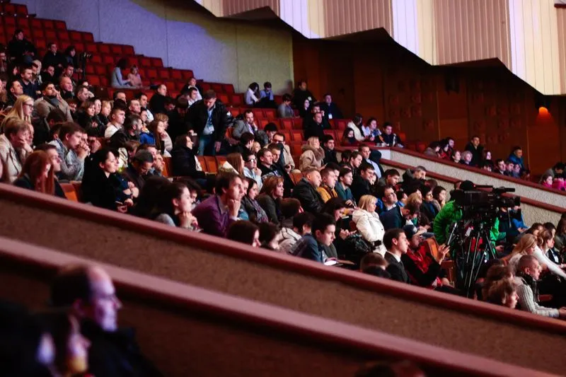 Public assis dans les gradins rouges d'une grande salle, avec un cadreur et sa caméra installés au milieu des rangs