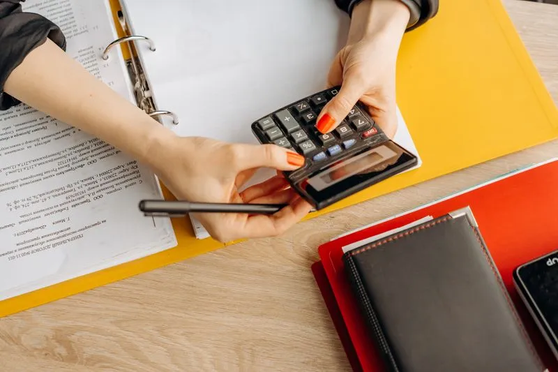 Mains d'une femme tenant une calculatrice et un stylo au-dessus d'un classeur ouvert rempli de documents