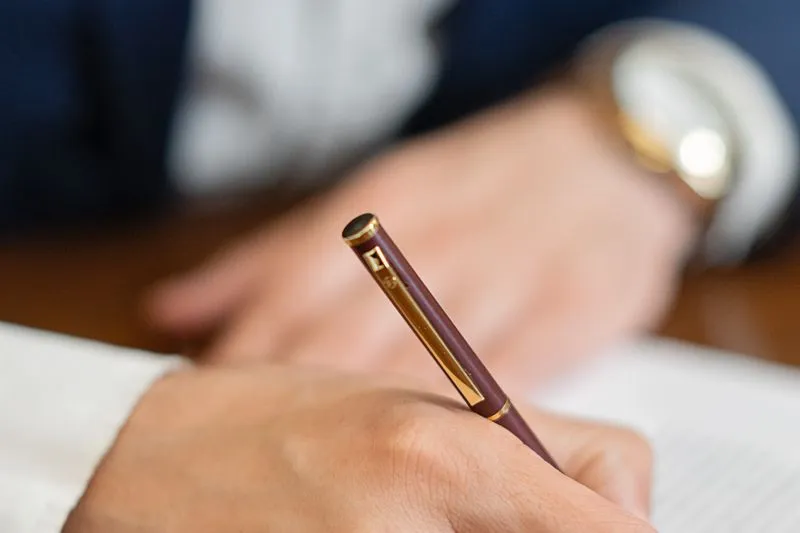 Main signant un document avec un stylo bordeaux et doré, une seconde personne floue à l'arrière-plan