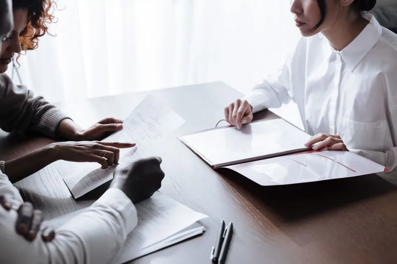 Couple assis face à une conseillère en chemise blanche, documents et dossier ouverts sur la table entre eux