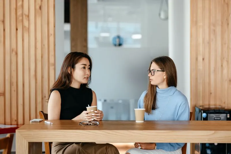 Deux jeunes femmes qui discutent face à face à une grande table en bois, un gobelet de café devant elles