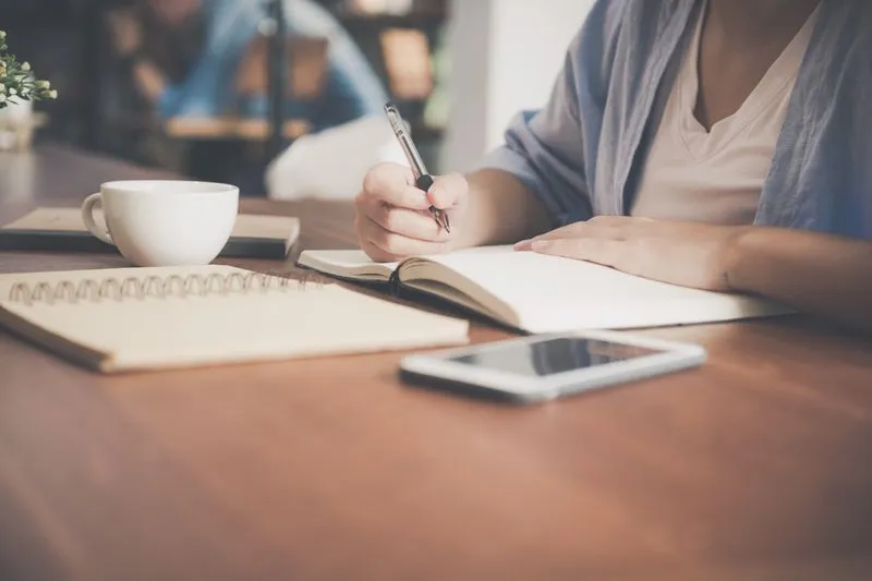 Main de femme qui écrit au stylo dans un carnet ouvert sur une table en bois, près d'une tasse blanche et d'un téléphone