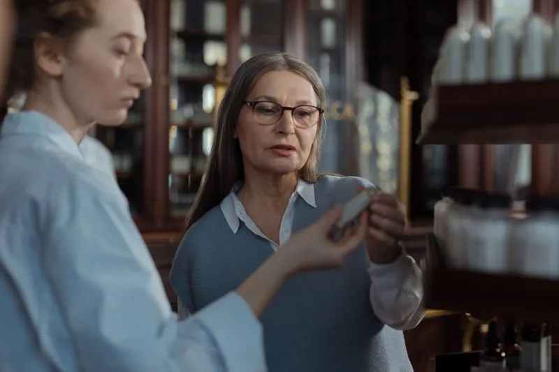 Une jeune femme en chemise blanche montre un petit flacon à une femme à lunettes, devant des étagères en bois chargées de flacons