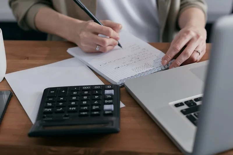 Main qui note des calculs dans un carnet à spirale, à côté d'une calculatrice et d'un ordinateur portable